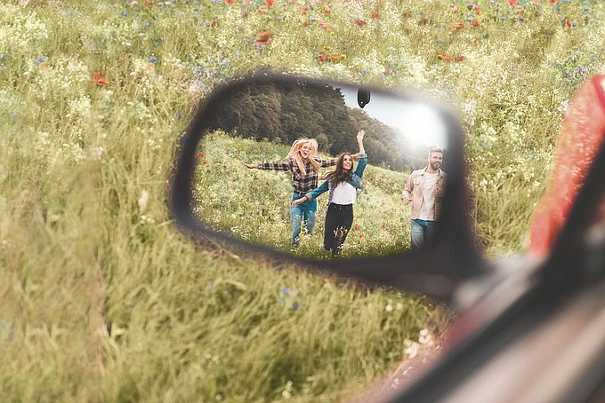 In einem Rückspiegel sind zwei Frauen und ein Mann zu sehen, die über eine Wiese laufen.