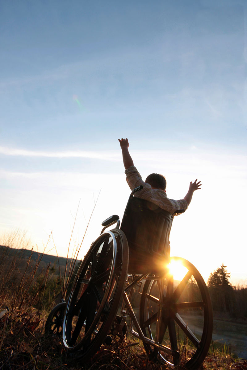 auf dem Foto hebt ein Rollstuhlfahrer, vor einem Sonnenuntergang sitzend, die Arme.