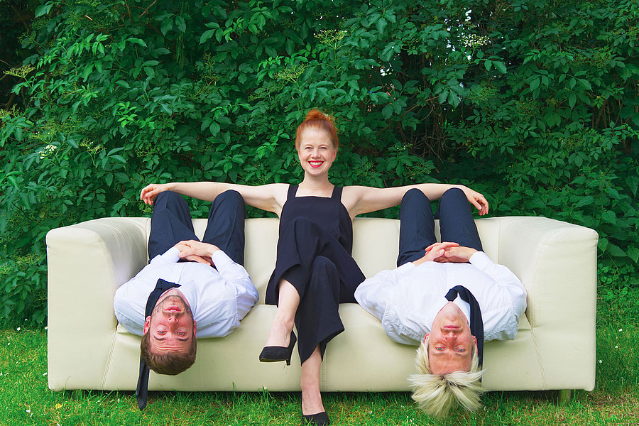 Eine Frau sitzt auf einer Couch. Links und rechts neben ihr liegt ein Mann auf dem Rücken.
