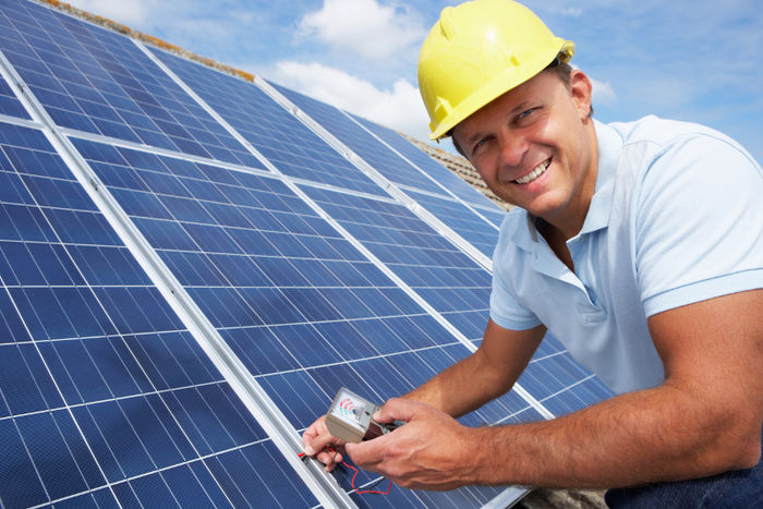 Das Foto zeigt einen Photovoltaikmonteur mit gelben Helm, der auf dem Dach sitzt und Solarpanele kontrolliert.
