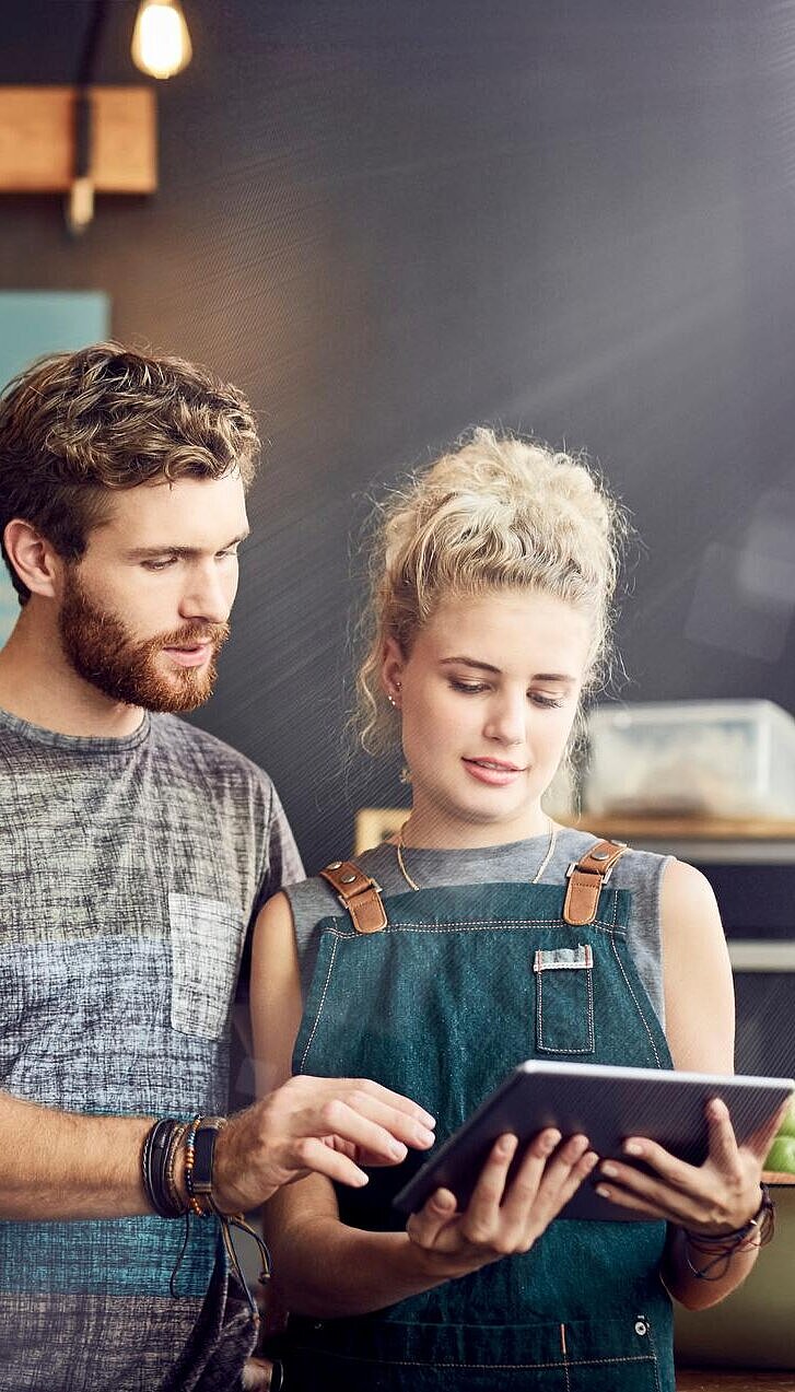Ein Mann und eine Frau schauen auf ein Tablet hinter der Theke in einem gastronomischen Betrieb, in dem sie arbeiten.