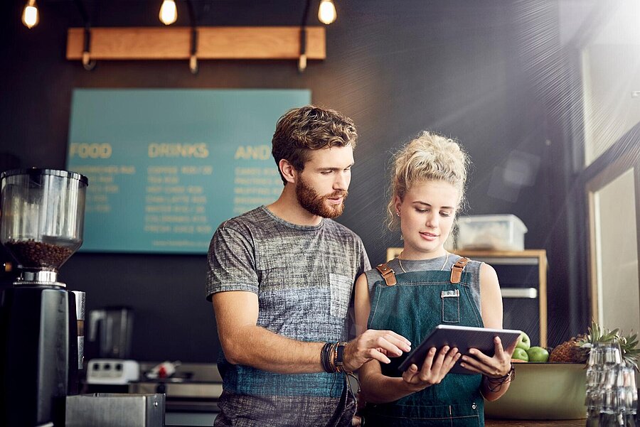 Ein Mann und eine Frau schauen auf ein Tablet hinter der Theke in einem gastronomischen Betrieb, in dem sie arbeiten.