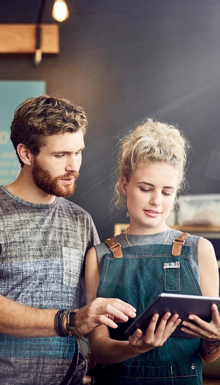 Ein Mann und eine Frau schauen auf ein Tablet hinter der Theke in einem gastronomischen Betrieb, in dem sie arbeiten.