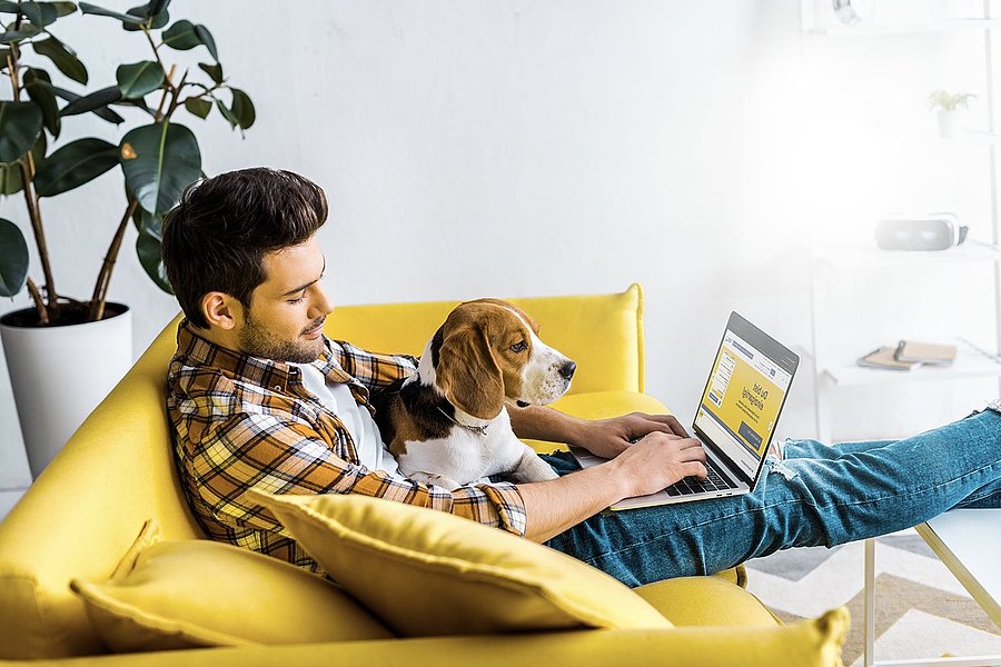 Ein Mann sitzt mit einem Hund auf einer Couch und schaut auf einen Laptop.