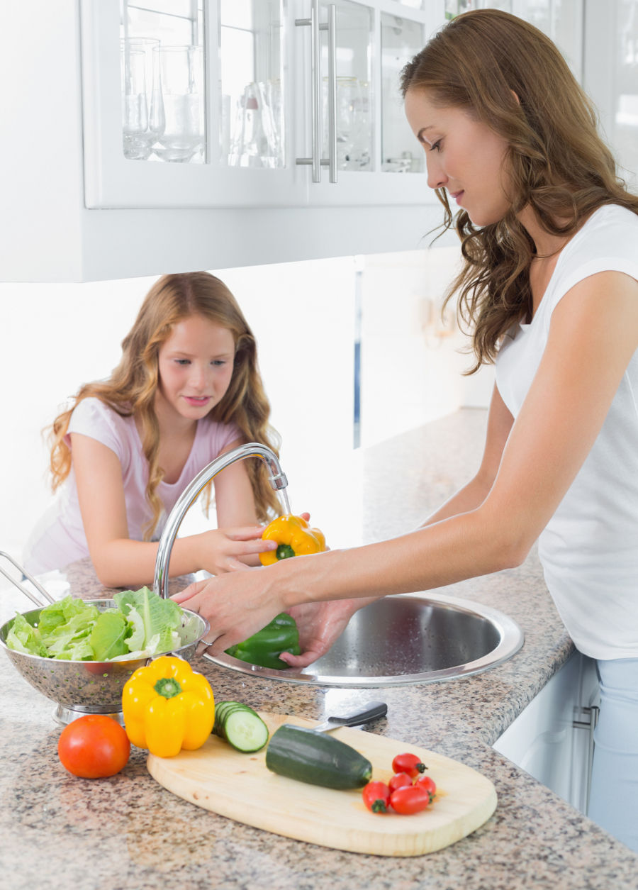 Das Foto zeigt eine Frau und ein Kind, die in der Küche stehen und verschiedene Gemüsearten unter dem Wasserhahn waschen.