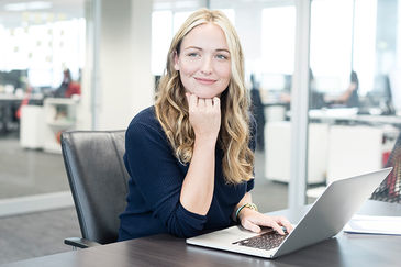 Das Foto zeigt eine junge Frau, die im Büro vor ihrem Laptop sitzt und in die Ferne schaut.