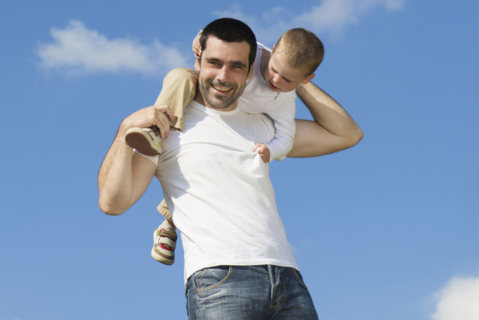 Ein junger Mann mit weißem T-shirt trägt einen kleinen Jungen auf seiner Schulter. Im Hintergrund ist blauer Himmel.